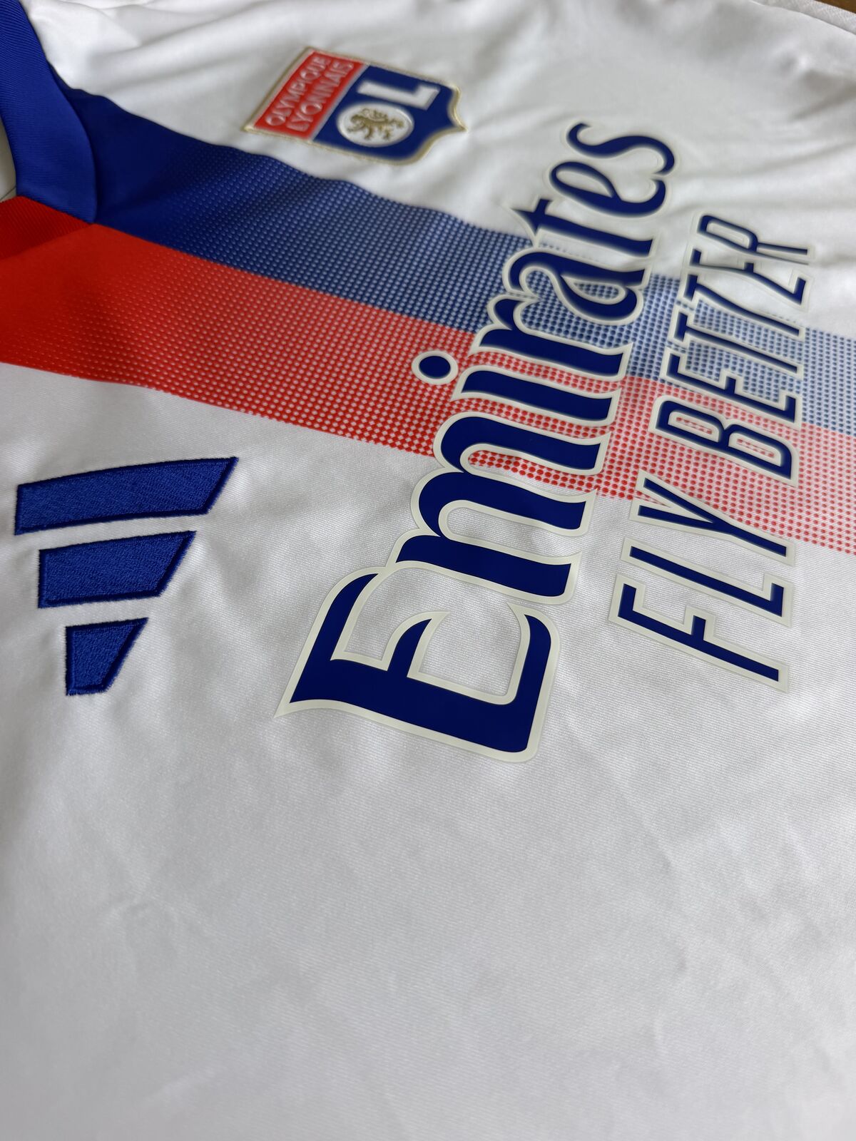 Angled close-up of the embroidered blue adidas logo and the applied Emirates Fly Better sponsor lettering