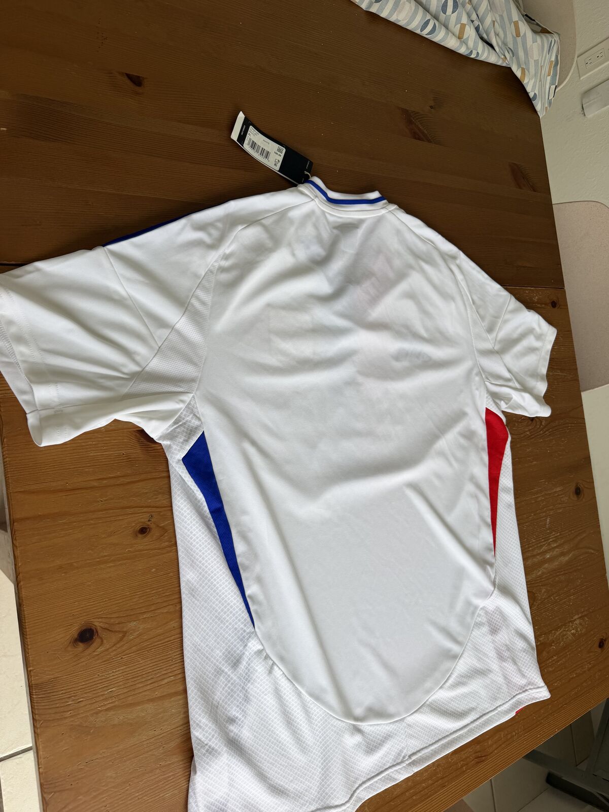 Back of the shirt laid flat with the cardboard hangtag still attached, showing plain white back with blue and red flank panels